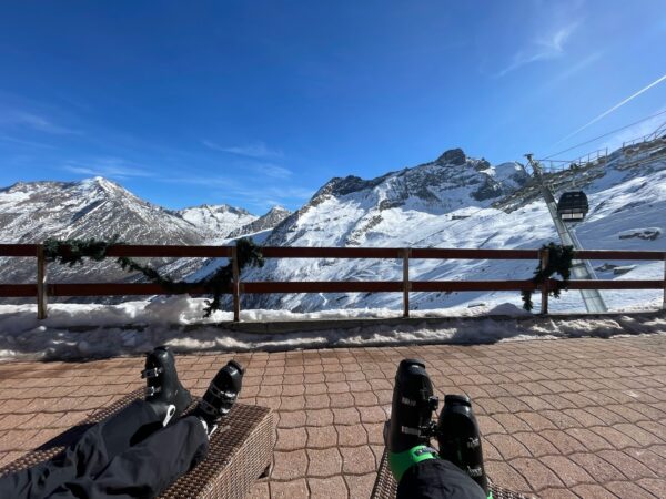 My legs wearing ski boots, reclining on a deck chair, with snowy swiss mountains in the distance. 